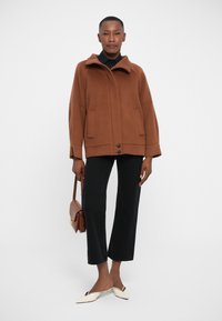 Cappotto di lana marrone con colletto alto, due tasche frontali e chiusura a doppio bottone. Abbinato a pantaloni neri e scarpe a punta crema.