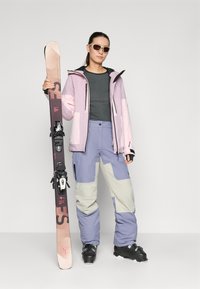 Skiantrekket har en lys rosa jakke med svarte detaljer, grå topp og lilla skibukser med grå lapper, og holder et par ski.