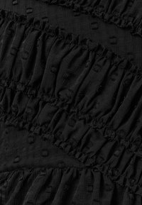 Tejido negro con un patrón texturizado que presenta cuadrados elevados; diseño con volantes y secciones fruncidas que crean líneas horizontales.