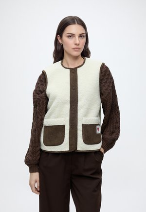 Femme portant un gilet en polaire blanc avec des poches en velours côtelé marron et des finitions sur un pull marron texturé à manches longues, debout devant un fond uni.