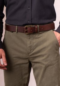 Brown leather belt with a textured surface, featuring a brass buckle. Worn over olive green pants, highlighting a casual style.