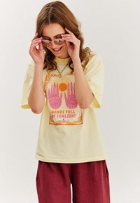 Žluté bavlněné tričko s růžovým grafickým designem rukou a slunce, text "RUKY PLNÉ SLUNEČNÍHO SVITU," v kombinaci s rezavě zbarvenými kalhotami.