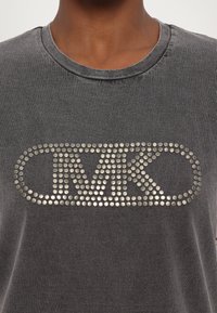 Camiseta de algodón gris con cuello redondo, que presenta un logo metálico con tachuelas "MK" en el pecho. Textura suave, ajuste casual.