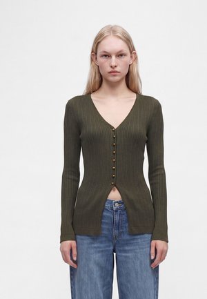 Junge Frau mit langen blonden Haaren, die eine olivgrüne, gerippte Strickjacke mit Knöpfen und blaue Jeans trägt, vor einem einfachen Hintergrund stehend.