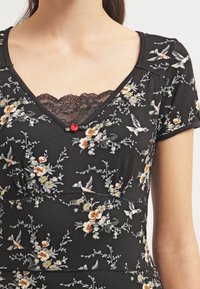 Svart blommig klänning med vita, orange och gula blommönster, spetskrage, korta ärmar och en dekorativ röd knapppynt.