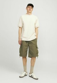 Kremowa koszulka z krótkim rękawem w połączeniu z oliwkowymi szortami cargo. Model nosi beżowe skarpetki oraz białe sneakersy z czarnymi akcentami. Prosty, casualowy strój.