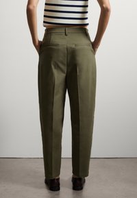 Pantalon olive ajusté, avec une texture lisse, des poches arrière et un pli sur le devant. Porté avec des chaussures sombres.