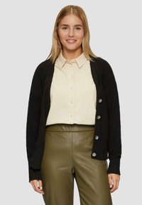 Schwarze Strickjacke mit einer Knopföffnung, getragen über einem hellen Hemd und olivgrünen Lederhosen. Weicher Stoff und lässige Passform.