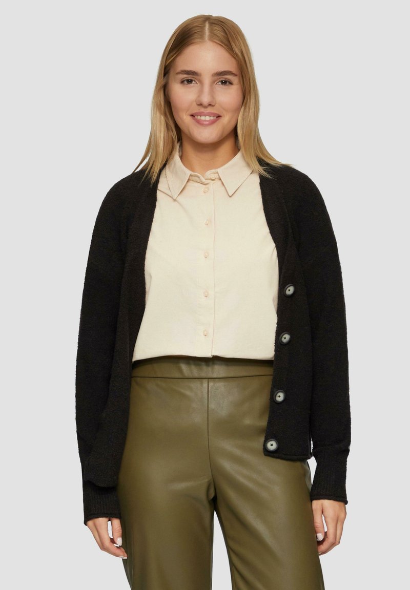Schwarze Strickjacke mit einer Knopföffnung, getragen über einem hellen Hemd und olivgrünen Lederhosen. Weicher Stoff und lässige Passform.