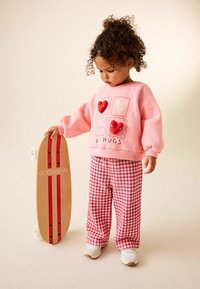 Bambino che indossa una felpa rosa con motivi a cuore e pantaloni rossi a quadri, tiene in mano uno skateboard di legno con strisce rosse. Scarpe bianche.