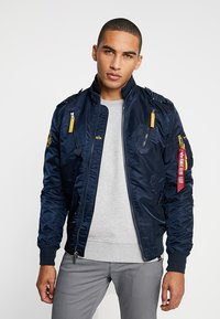 Navy bomber jacket with zippers and pockets, featuring yellow accents and patches. Smooth, shiny fabric; ribbed cuffs and collar.