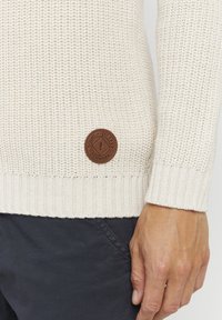 Valkoisen villapaidan neule, jossa resorit hihansuissa ja helmassa. Alemmassa sivussa pyöreä ruskea nahkalogo, jossa brändin nimi ja vuosiluku.