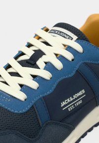 Μπλε αθλητικό παπούτσι με συνδυασμό υφασμάτων με υφή, λευκά κορδόνια και ετικέτα στο πλάι που αναγράφει "JACK&JONES EST.1990".