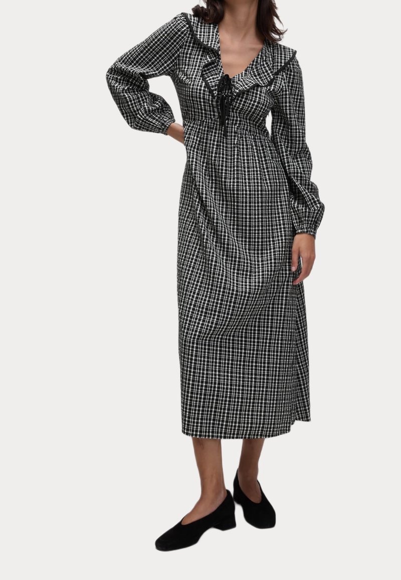 Femme portant une robe midi à manches longues à carreaux noirs et blancs avec un col à volants et des chaussures noires à talons bas.