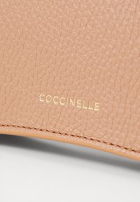 Struktūrinė persikų oda su auksine "COCCINELLE" logotipo detale. Lygaus paviršiaus kraštai ir siūlės matomos palei siūles.