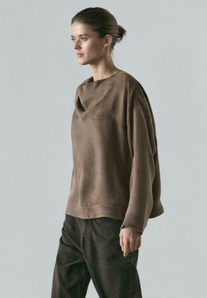 Vrouw met haar in een knot, draagt een loszittend lang bruin shirt en donkere broek, staand tegen een effen lichtgrijze achtergrond.