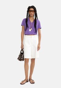 Fioletowa koszulka z dekoltem w serek, białe szorty do kolan, brązowa torebka w kropki, brązowe klapki i długie, plecione włosy z akcesoriami.