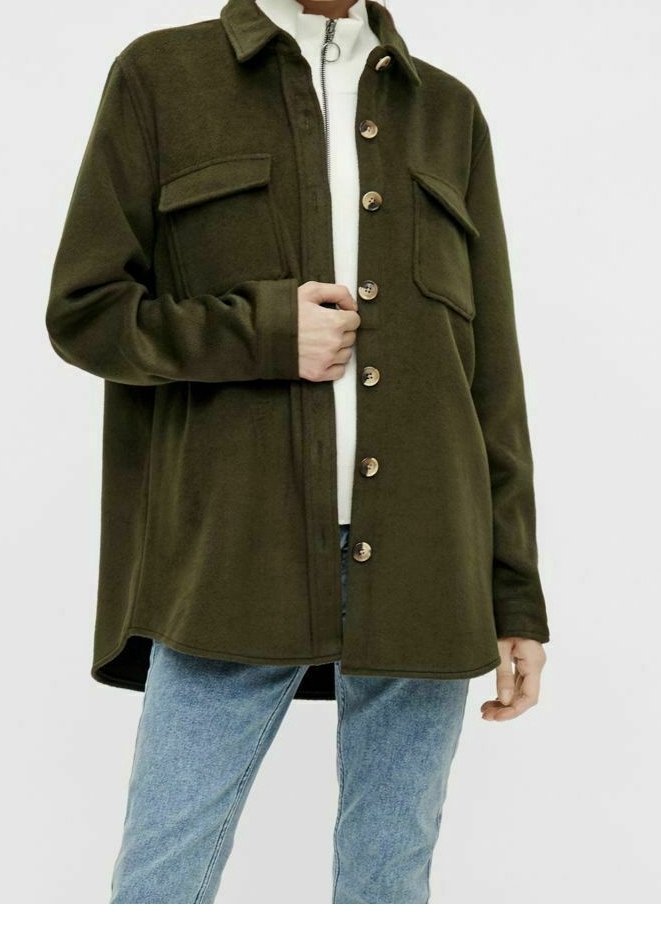 Personne portant une veste boutonnée vert olive avec des poches à rabat sur la poitrine, un haut blanc et un jean bleu clair, main tenant la veste près de la taille.