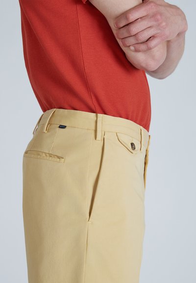Chinos amarillos con un corte ajustado, que cuentan con una parte delantera plana, bolsillos laterales y traseros, y un detalle de botón marrón en la cintura. Llevados con una camisa roja.