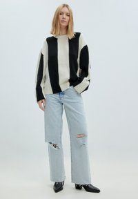 Übergroßer Pullover mit schwarz-weißen Streifen, geripptem Ausschnitt; kombiniert mit hellblauen, ausgewaschenen Schlagjeans und schwarzen Knöchelschuhen.