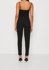 Mono negro con piernas ajustadas y entalladas, tirantes delgados ajustables y un cuerpo texturizado. Cuenta con una cremallera en la parte posterior y un tejido suave.