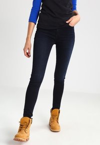 Mörkblå slim fit jeans med en jämn textur, bärs tillsammans med en orange-brun arbetskänga. Modellen står upprätt.