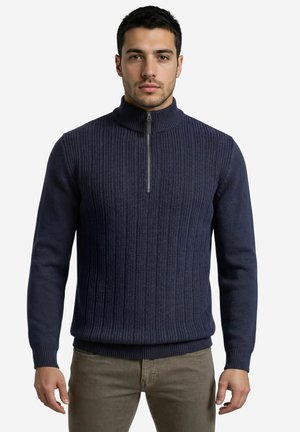 Mann, der nach vorne blickt und einen marineblauen, gerippten Pullover mit halbem Reißverschluss sowie olivgrüne Hosen trägt, vor einem schlichten hellen Hintergrund.