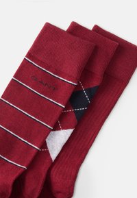 Three pairs of red socks featuring a solid color, horizontal stripes, and a diamond pattern in navy and white. Soft knit texture.
