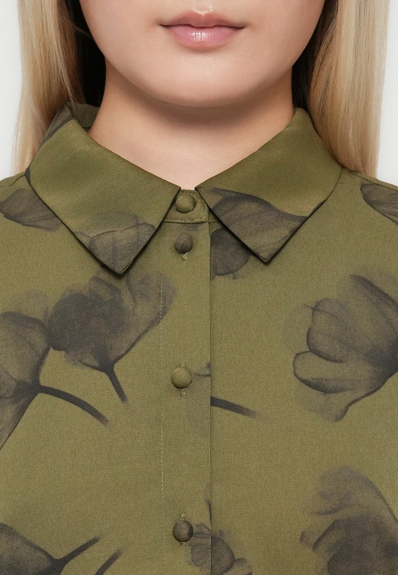 Nahaufnahme des unteren Gesichts und Halses einer Frau, die ein olivgrünes, mit schwarzen Blumenmustern und stoffbezogenen Knöpfen versehenes Hemd mit Kragen trägt.