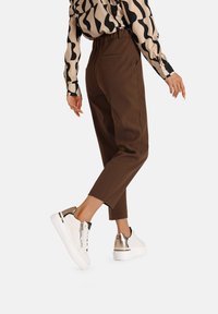 Pantaloni tapered marroni con vita alta, abbinati a sneaker bianche con dettagli dorati e una parte superiore testurizzata. Blusa a fantasia visibile.