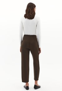 Donna con capelli castani fino alle spalle, che indossa un top bianco a maniche lunghe, pantaloni alti marrone scuro e ballerine nere, vista da dietro.