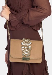 Beżowa kopertówka z paskiem z złotym łańcuszkiem i ozdobami z biżuterii na klapie; wykonana z gładkiego materiału o połyskującym wykończeniu.
