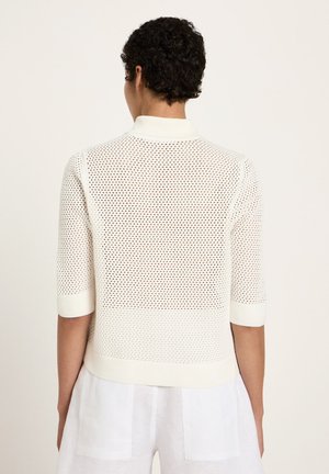 Person mit kurzen lockigen Haaren, trägt ein weißes, perforiertes Strickoberteil und weiße Hosen, steht mit dem Rücken zum Betrachter vor einem schlichten Hintergrund.