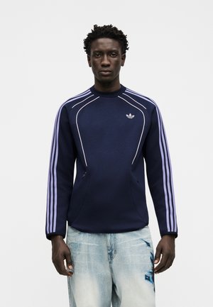 Mężczyzna ubrany w granatową bluzę Adidas z liliowymi paskami na rękawach oraz jasnoniebieskie dżinsy z ciemnoniebieskim nadrukiem na lewym udzie.