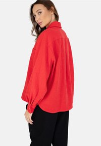 Czerwona, oversize koszula z kołnierzykiem, długimi balonowymi rękawami i zaokrąglonym dołem. Teksturowana tkanina z jedną kieszonką na klatce piersiowej. Połączona z czarnymi spodniami.