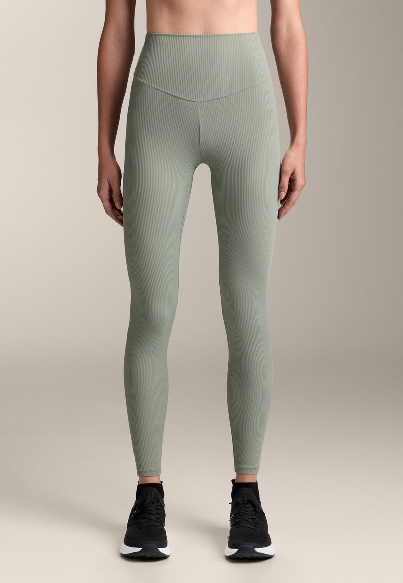 Magas derekú, bordázott leggings puha zsályazöld színben, sima felülettel. Fekete sportcipővel párosítva, texturált talppal.