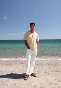 Άντρας που στέκεται σε αμμώδη παραλία με τα χέρια στις τσέπες, φορώντας πουκάμισο με κοντά μανίκια και σχέδια, λευκό παντελόνι και καφέ σανδάλια δίπλα στον ωκεανό.