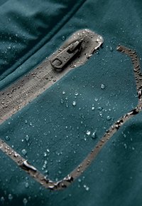 Wasserabweisende Jacke in tiefem Türkis, mit einem grauen Reißverschluss, Tropfen auf dem Stoff und einem schlanken Design mit einem minimalistischen Logo-Akzent.