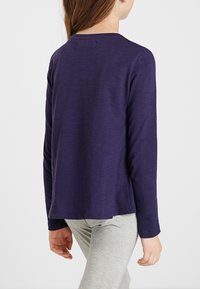 Dítě oblečené v ponořené dlouhé rukávy tmavě modré košile a světle šedých kalhotách, viděné zadečku na jednolitém pozadí.