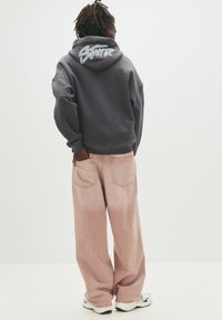 Grå hoodie med et signaturlogo på ryggen, parret med løse lyse rosa jeans. Sættet suppleres af hvide sneakers.