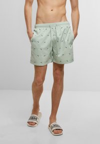 Svijetlozelene kupaće gaće s crnim uzorkom riba, elastična su viša i imaju funkcionalne bočne džepove. Uparene su sa bež laganim japankama s natpisom.