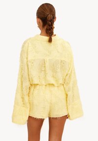 Mujer con cabello trenzado vestida con una camiseta amarilla de manga larga texturizada de gran tamaño y pantalones cortos a juego, de pie con la espalda hacia adelante.