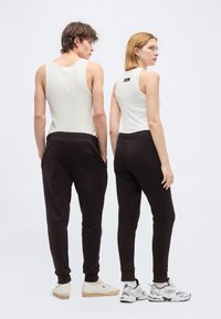 Δύο άτομα με λευκές αμάνικες μπλούζες και μαύρα παντελόνια τζόγκινγκ, στεκόμενα πλάτη με πλάτη μπροστά σε ένα απλό λευκό φόντο.