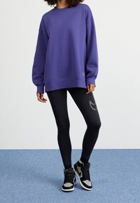 Sudadera oversized púrpura con mangas largas, combinada con leggins negros que tienen un acento dorado. La modelo lleva zapatillas altas negras y grises.