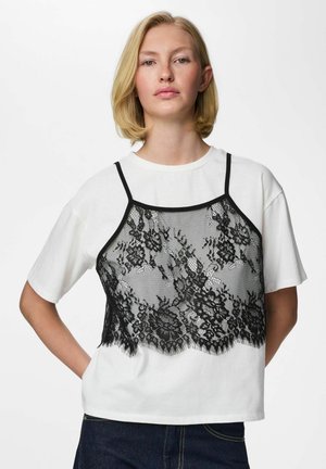 Femme blonde portant une chemise blanche à manches courtes avec un caraco en dentelle noire à motifs floraux superposé, debout devant un fond blanc.