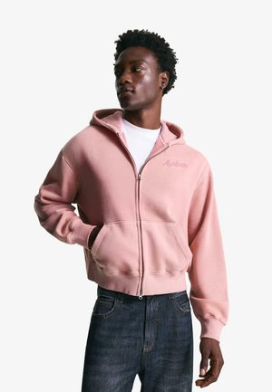 Pink zip-up hoodie made of soft fabric with a front pocket, ribbed cuffs, and embroidered branding on the chest. Paired with blue jeans.