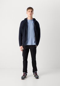 Felpa con zip blu navy sopra una maglietta azzurra chiara, pantaloni neri e sneakers multicolori con un design spesso e decorazioni a pattern.