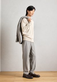 Pulover bej tricotat cu tiv cu nervuri, jachetă de costum gri aruncată pe umăr, pantaloni gri făcuți pe comandă și mocasini maro închis pe podea din lemn.