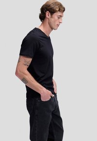 Fiatal férfi göndör hajjal és tetoválásokkal, fekete V-nyakú pólót és fekete farmert visel, oldalra fordulva, kezét a zsebében tartva.