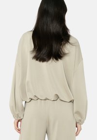 Top lungo a maniche lunghe in morbido tessuto beige, con vita e polsini elasticizzati, dalla texture liscia e silhouette rilassata.